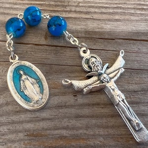 May include: A silver rosary with a blue and white enamel pendant featuring the Virgin Mary and a silver crucifix with a figure of Jesus Christ. The rosary beads are blue with white marbling.