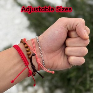 Puede incluir: Una mano con varias pulseras ajustables. Las pulseras incluyen una pulsera de cord&oacute;n trenzado rojo, una pulsera de cuentas de madera, una pulsera de cord&oacute;n rosa y una pulsera de cadena plateada. El texto "Adjustable Sizes" est&aacute; en rojo.