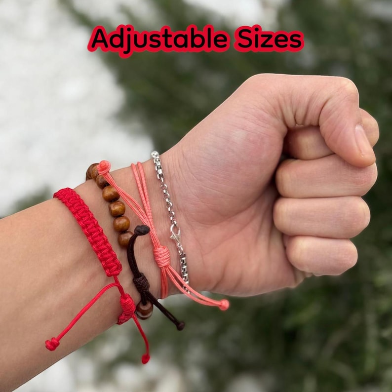 Puede incluir: Una mano con varias pulseras ajustables. Las pulseras incluyen una pulsera de cord&oacute;n trenzado rojo, una pulsera de cuentas de madera, una pulsera de cord&oacute;n rosa y una pulsera de cadena plateada. El texto "Adjustable Sizes" est&aacute; en rojo.