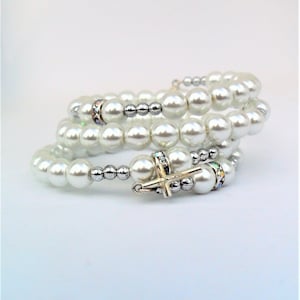 Puede incluir: Un brazalete de perlas blancas con un dije de cruz de plata y cristal. El brazalete está hecho de múltiples hileras de perlas y cuentas de plata.