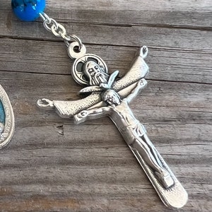 May include: Silver rosary with blue beads and a silver crucifix featuring a depiction of Jesus Christ on the cross. The crucifix has a detailed design with a halo around Jesus' head.