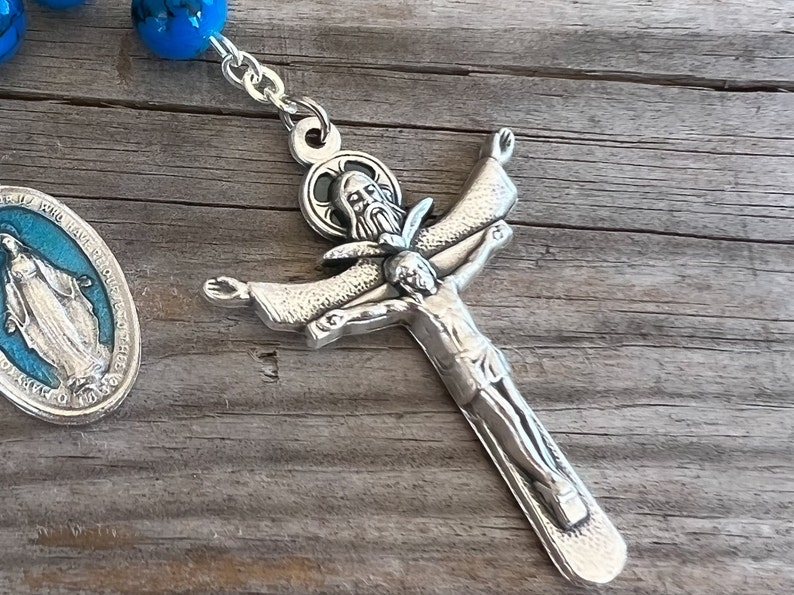 May include: Silver rosary with blue beads and a silver crucifix featuring a depiction of Jesus Christ on the cross. The crucifix has a detailed design with a halo around Jesus' head.