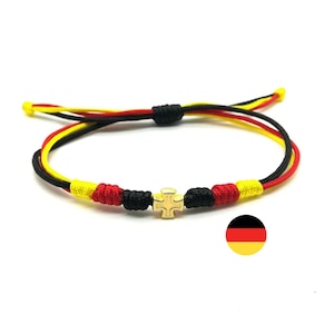 May include: A black, red, and yellow braided bracelet with a gold cross charm. The bracelet is tied with a knot and has a small German flag charm.