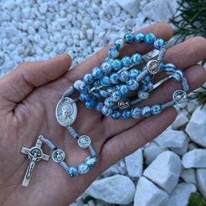 Könnte beinhalten: Ein Rosenkranz mit irisierenden blau-weißen Perlen. Der Rosenkranz hat ein silbernes Kruzifix und mehrere silberne Medaillons. Die Perlen sind auf einer grauen Schnur aufgefädelt. Ein religiöser Gegenstand.
