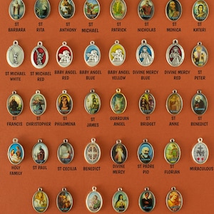 May include: A collection of 36 oval-shaped metal charms with various religious figures and images. The charms are silver, gold, and red with colorful images of saints, angels, and religious symbols. The charms are arranged in rows on a brown background.