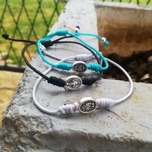 Puede incluir: Tres pulseras trenzadas con dijes de plata. Las pulseras son blancas, negras y turquesa. Los dijes presentan una imagen religiosa.