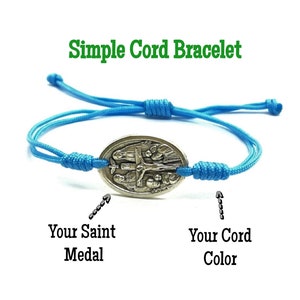 May include: A simple cord bracelet with a silver oval charm featuring a religious image. The bracelet is blue and the charm is silver. The text "Your Saint Medal" and "Your Cord Color" is written below the bracelet.