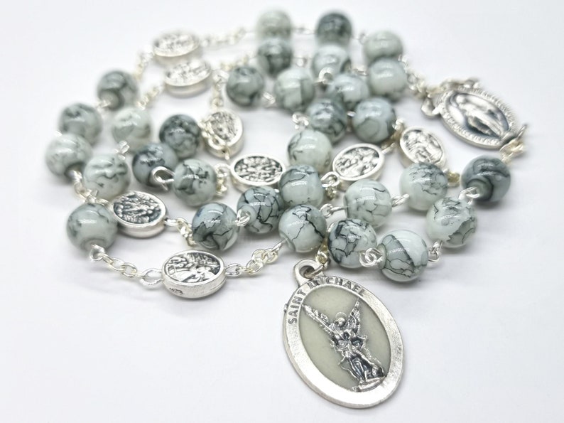 May include: A white and black marbled rosary with silver beads and a silver pendant featuring Saint Michael the Archangel. The pendant has the words "Saint Michael" inscribed on it.