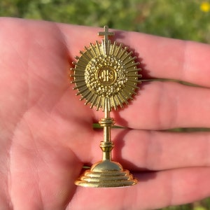 Puede incluir: Un ostensorio de metal dorado con una cruz en la parte superior y una base circular. El ostensorio tiene un diseño de rayos de sol y las letras "IHS" en el centro.