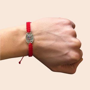 Puede incluir: Pulsera roja tejida con un dije ovalado plateado que representa una figura religiosa. La pulsera es ajustable con un cierre de nudo corredizo. El dije está centrado en la banda roja tejida.