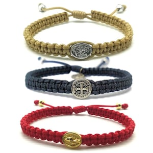 May include: Three braided bracelets with different colored cords and metal charms. The first bracelet has a beige cord and a silver charm with a religious image. The second bracelet has a navy blue cord and a silver charm with a cross. The third bracelet has a red cord and a gold charm with a religious image.