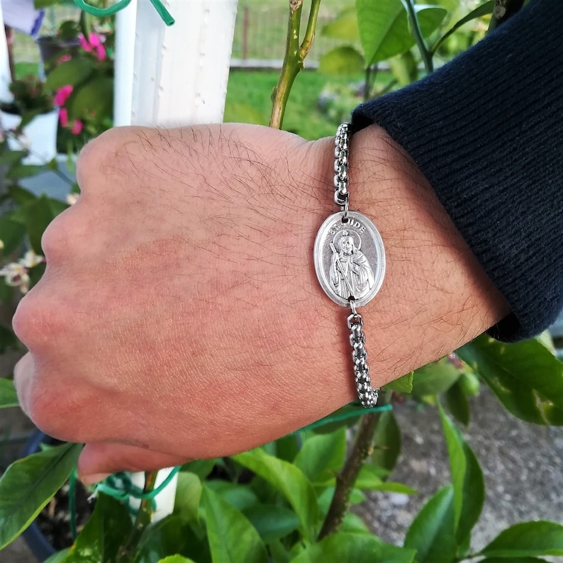 Pu&ograve; includere: Un braccialetto a catena d'argento con un ciondolo rotondo d'argento che presenta una figura religiosa. Il ciondolo ha la scritta "Jude" incisa.