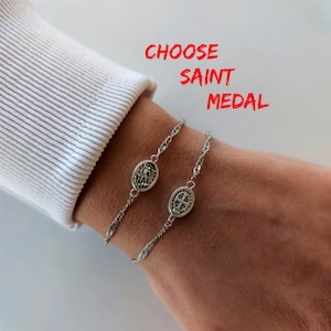 Op de afbeelding: Twee zilveren kettingarmbanden met ovale medaillons. De medaillons tonen religieuze afbeeldingen. De tekst "CHOOSE SAINT MEDAL" wordt boven de armbanden weergegeven.
