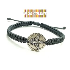 May include: Grey braided bracelet with a silver charm featuring a dove with a halo. The charm is round and has a scalloped edge. The text "IN 25 COLORS" is visible above the bracelet.