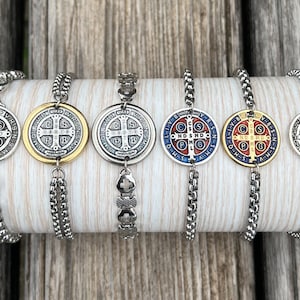 Puede incluir: Seis pulseras de cadena plateada con un colgante redondo que presenta una medalla de San Benito. Las medallas tienen una cruz en el centro y están decoradas con esmalte dorado, azul y rojo.