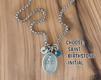 May include: Silver-toned necklace with a detailed pendant of a saint, a small initial charm, and a blue gemstone. The chain has a textured design. Text on the image reads "CHOOSE: -SAINT -BIRTHSTONE -INITIAL".