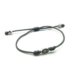 May include: A gray cord bracelet with a silver oval charm featuring a black and white design. The charm has a knot on each side.