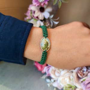 May include: A green braided bracelet with a silver oval charm featuring a religious image. The charm has a white background with a colorful image of a person in a green robe.