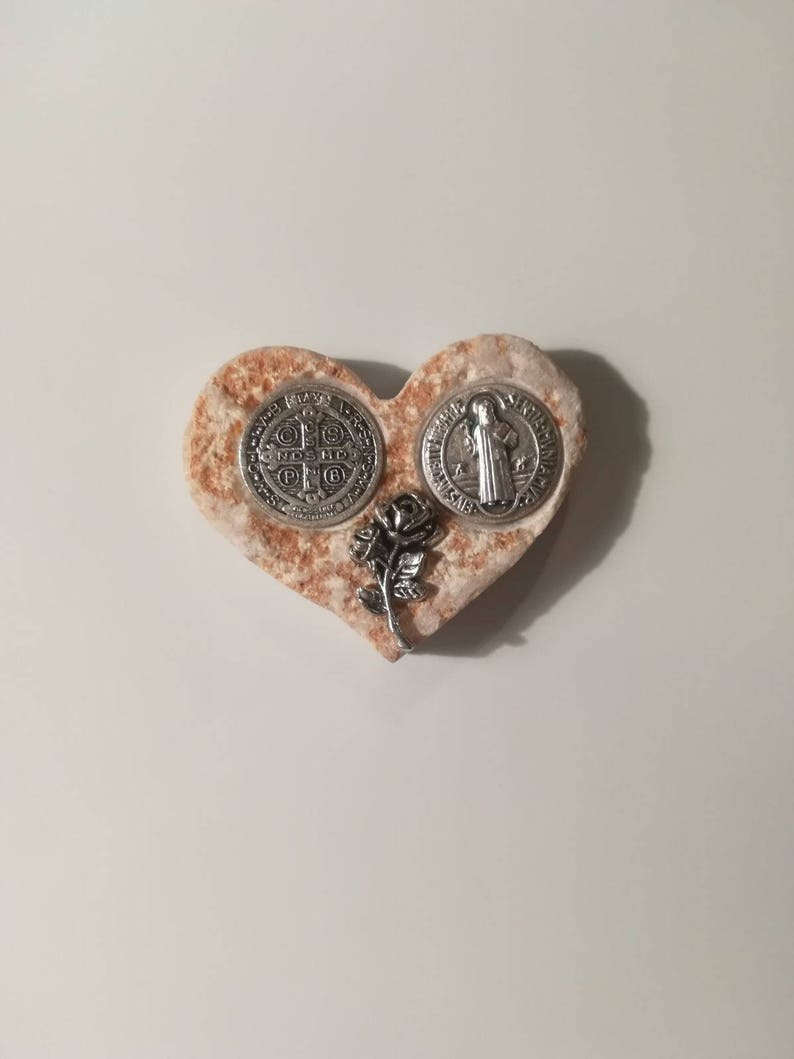K&ouml;nnte beinhalten: Ein herzf&ouml;rmiger Stein mit einer silbernen St. Christopher-Medaille und einer silbernen St. Benedikt-Medaille sowie einer silbernen Rose.