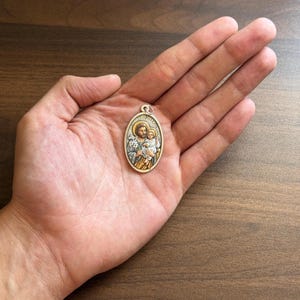 Puede incluir: Un colgante de metal de forma ovalada con un relieve coloreado de San José sosteniendo al niño Jesús. El diseño incluye un halo y detalles en tono dorado, con el texto ST. JOSEPH grabado a lo largo del borde superior del colgante.