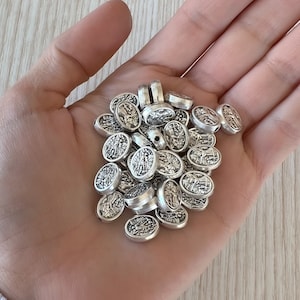 May include: A pile of silver oval-shaped beads with intricate designs. The beads have a slightly textured surface and are perfect for jewelry making or crafting.
