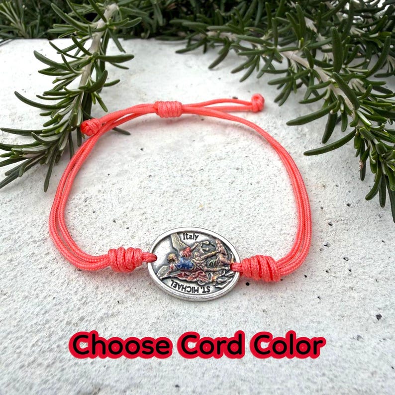 Puede incluir: Una pulsera de color coral con un colgante ovalado plateado que presenta una imagen de San Miguel y la palabra "Italia". La pulsera est&aacute; hecha de cord&oacute;n tejido y tiene nudos ajustables. El texto "Choose Cord Color" es rojo.