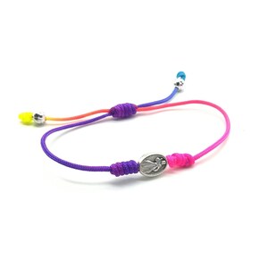 May include: A colorful braided bracelet with a silver charm featuring a religious figure. The bracelet is made with multiple strands of cord in shades of yellow, orange, red, purple, and pink.