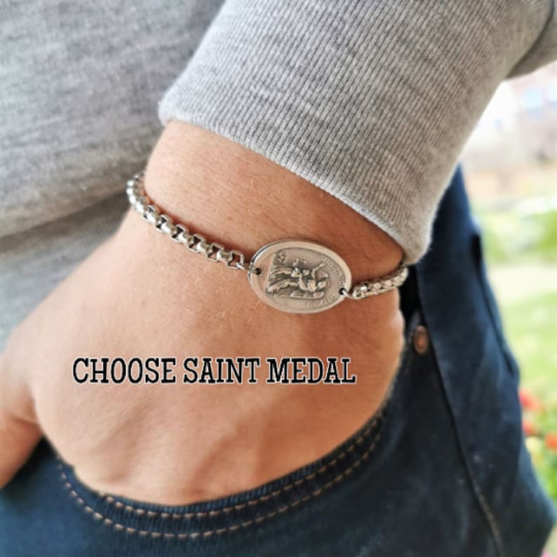 Pu&ograve; includere: Un braccialetto a catena d'argento con un ciondolo ovale d'argento che presenta una figura religiosa. Il testo "CHOOSE SAINT MEDAL" &egrave; visibile sotto il braccialetto.