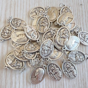 Puede incluir: Un montón de dijes ovalados de plata con la imagen de San Cristóbal y el texto "St. Christopher" en cada dije.