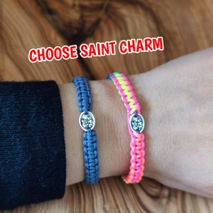 Può includere: Due braccialetti con un designo intrecciato colorato e ciondoli d'argento. Il testo "CHOOSE SAINT CHARM" è visualizzato sopra i braccialetti.