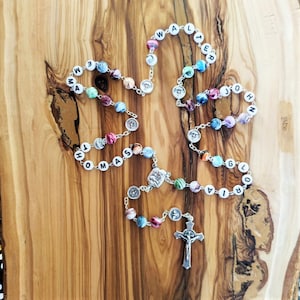 May include: A colourful beaded rosary with silver accents. The beads are made of glass and have a variety of colours, including blue, green, pink, and purple. The rosary is strung on a silver chain and has a silver crucifix at the end. The beads are inscribed with the names "Walter", "John", "Olivia", "Thomas", and "Emma".