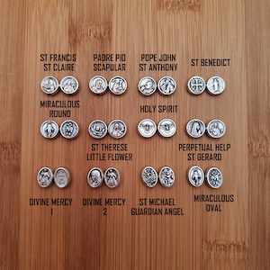 May include: A collection of silver-toned metal medallions featuring various religious figures and symbols. The medallions are arranged in rows on a wooden surface. The medallions include images of St. Francis, Padre Pio, Pope John, St. Anthony, St. Benedict, the Miraculous Round, the Holy Spirit, St. Therese, Perpetual Help, St. Gerard, Divine Mercy, St. Michael, and the Miraculous Oval.