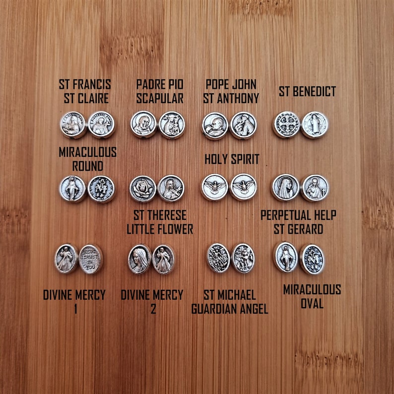 May include: A collection of silver-toned metal medallions featuring various religious figures and symbols. The medallions are arranged in rows on a wooden surface. The medallions include images of St. Francis, Padre Pio, Pope John, St. Anthony, St. Benedict, the Miraculous Round, the Holy Spirit, St. Therese, Perpetual Help, St. Gerard, Divine Mercy, St. Michael, and the Miraculous Oval.