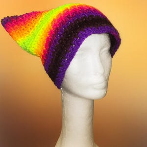 Può includere: Un cappello all'uncinetto fatto a mano con una punta appuntita, che mostra una sfumatura arcobaleno di colori. Il cappello presenta verde neon, arancione, rosa e viola. È esposto su una testa di manichino bianca.