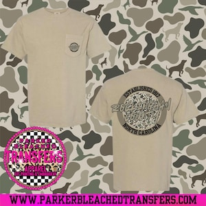 May include: A tan short-sleeved t-shirt with a pocket and a graphic on the back. The back graphic reads "Rockingham County North Carolina" in a circular design. The shirt also has the text "Parker Bleached Transfers" on the front.