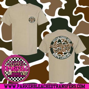 Könnte beinhalten: Beiges T-Shirt mit einer Tasche, die ein kreisförmiges Design mit Text und einem Camouflagemuster zeigt. Auf der Rückseite des Shirts befindet sich ein größeres, passendes Design mit dem Text "Rockingham County, North Carolina". Kurzärmlig.