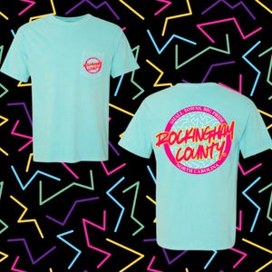 May include: Two light blue t-shirts featuring the text "Rockingham County" in a pink and white design. One shirt has the design on the front pocket, and the other has the design on the back. The shirts are set against a black background with colourful geometric shapes.