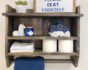 May include: A rustic wooden bathroom shelf with three shelves, a towel bar, and a sign that reads "PLEASE SEAT YOURSELF". The shelf is decorated with a white unicorn figurine, a blue glass jar, white towels, blue towels, and two rolls of toilet paper.
