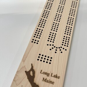 May include: A wooden cribbage board with a custom design featuring the outline of Long Lake, Maine. The board has a compass and the text "Long Lake Maine" engraved on it.