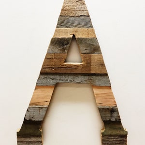 May include: A large wooden letter A made from reclaimed wood planks. The letter is painted in shades of brown, gray, and green. The letter is standing upright on a white background.
