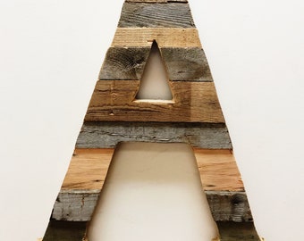 May include: A large wooden letter A made from reclaimed wood planks. The letter is painted in shades of brown, gray, and green. The letter is standing upright on a white background.