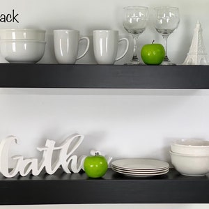May include: Two black floating shelves with white ceramic bowls, mugs, and plates. The top shelf also has two clear wine glasses and a green apple. The bottom shelf has a white wooden sign that says "Gather" and a green apple.