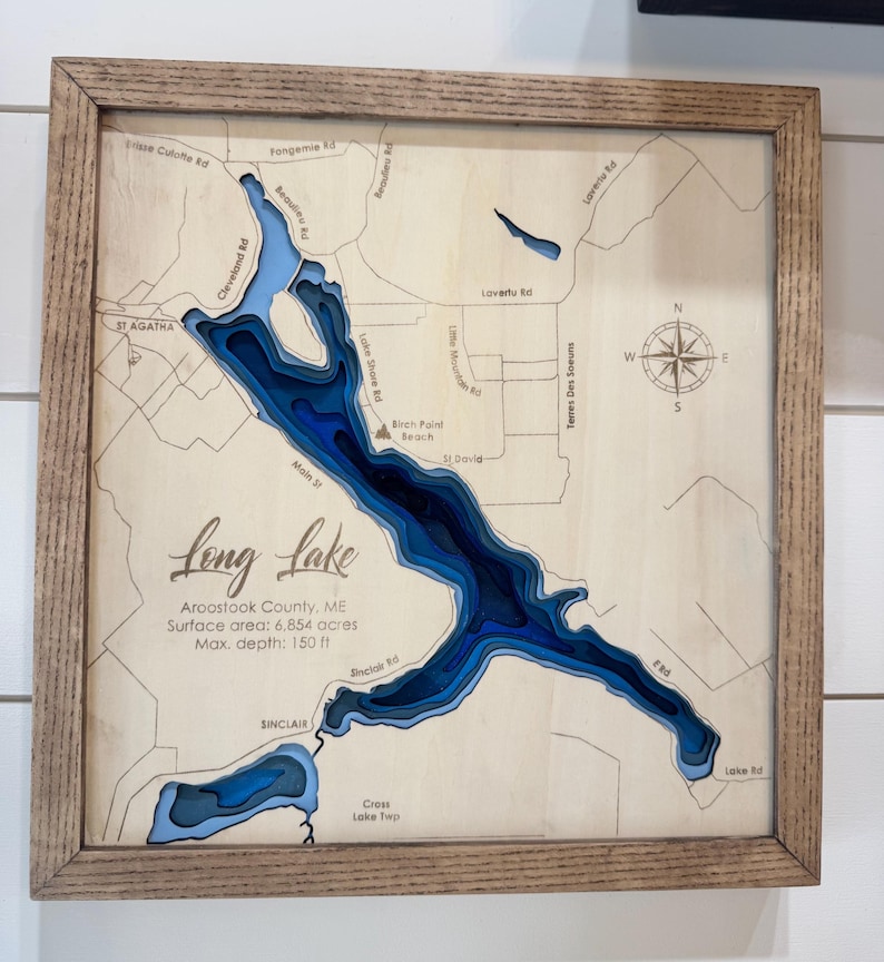 Puede incluir: Un mapa de madera enmarcado del lago Long en el condado de Aroostook, Maine. El mapa presenta un dise&ntilde;o en capas detallado que muestra la profundidad del lago en tonos de azul. El texto incluye el nombre del lago, la superficie y la profundidad m&aacute;xima de 45,72 metros.