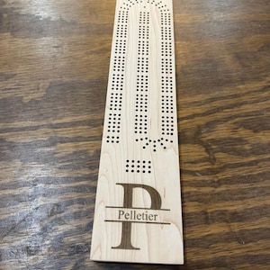 May include: A wooden cribbage board with a personalized initial "P" and the name "Pelletier" engraved on the bottom. The board has a traditional design with a curved track and holes for pegs.