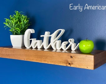 Puede incluir: Una estantería de madera con un tinte marrón claro. La estantería está decorada con una planta blanca en una maceta blanca, un letrero de madera blanco que dice "gather" y una manzana verde.