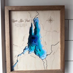 Puede incluir: Un mapa de madera enmarcado de Upper Shin Pond en el condado de Penobscot, ME. El mapa muestra la profundidad del estanque en capas de azul, con una superficie de 589 acres y una profundidad m&aacute;xima de 17,4 m. El marco es de color marr&oacute;n claro.