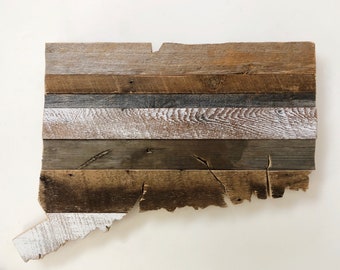 Puede incluir: Una rústica pieza de arte mural de madera con la forma del estado de Connecticut. La obra de arte está construida con tablones horizontales de madera reciclada en varios tonos de marrón, gris y blanco, creando una apariencia texturizada y envejecida. La pieza está montada sobre un fondo blanco.
