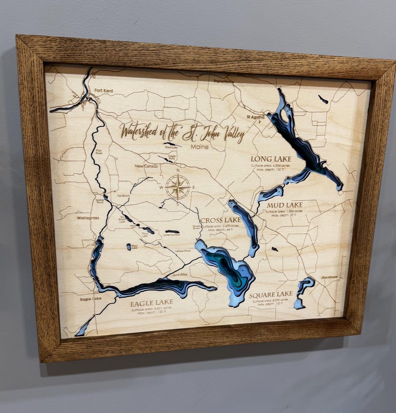Puede incluir: Mapa enmarcado de madera de la cuenca del valle de St. John en Maine. El mapa presenta capas cortadas con l&aacute;ser que detallan los lagos y la topograf&iacute;a, con etiquetas para lugares como Long Lake y Eagle Lake. El marco es de color marr&oacute;n c&aacute;lido.