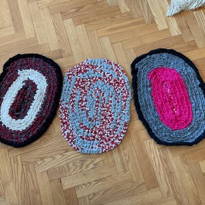 May include: Three oval-shaped braided rugs with different color combinations. The first rug is black with white and red stripes. The second rug is gray with white and red stripes. The third rug is gray with pink and white stripes.