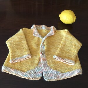 May include: A yellow knitted cardigan with a white and orange trim. The cardigan has a button closure and long sleeves.